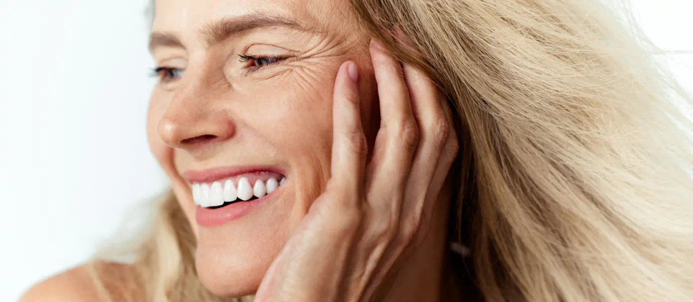 Joyful older woman with blonde hair touching her face, showing smooth skin and wrinkles